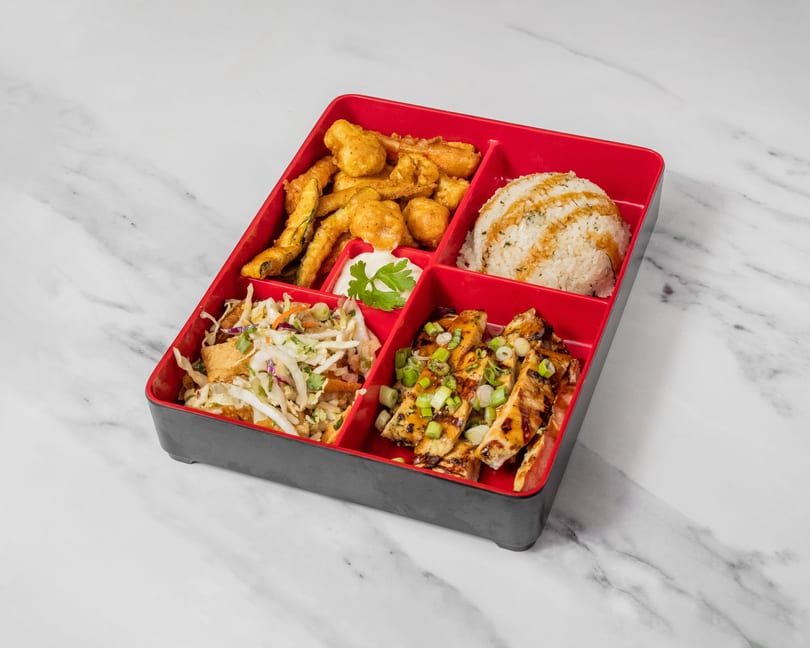 A bento box with fried zucchini, rice, coleslaw, and grilled chicken on a marble surface.