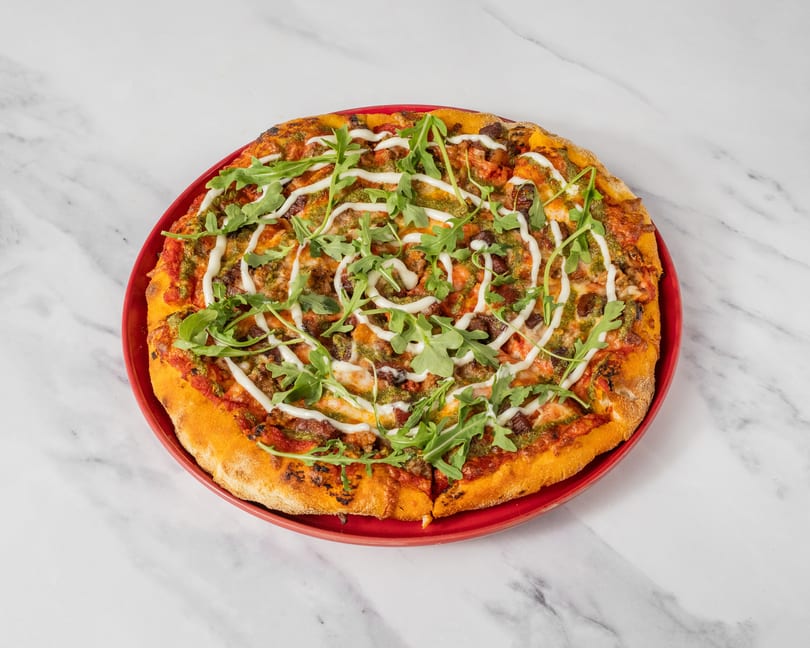 A whole pizza with pesto, tomato sauce, arugula, and a white sauce drizzle on a red plate.