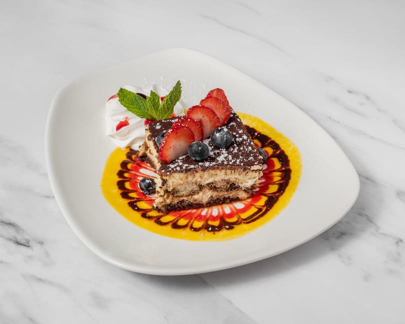 Tiramisu slice with strawberries, blueberries, whipped cream, and decorative sauce on a white plate.