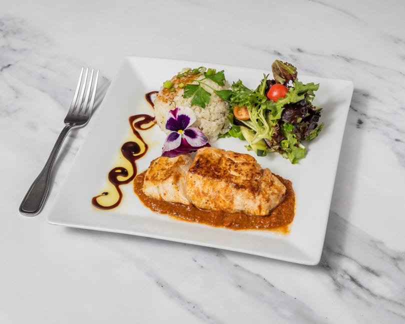 Pan-seared fish with rice, salad, and a purple edible flower on a white plate.
